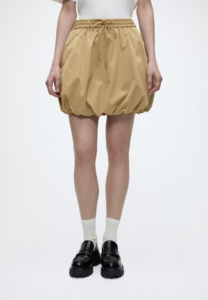 Jupe à pois beige avec taille élastique et cordon de serrage, portée avec des chaussettes blanches et des mocassins noirs épais sur une personne debout.