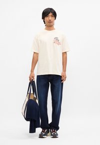 Κρέμα βαμβακερό t-shirt με γραφική εκτύπωση, συνδυασμένο με σκούρες τζιν παντελόνες. Το μοντέλο κρατάει μία ναυτική τσάντα και ένα πλεκτό πουλόβερ. Τα αθλητικά ολοκληρώνουν την εμφάνιση.