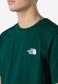 Zelené bavlněné tričko s kulatým výstřihem a krátkým rukávem, s bílým logem na levé straně hrudi. Hladká textura, ležérní design.