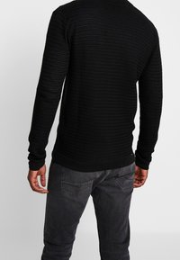 Man i en texturerad svart långärmad tröja och mörkgrå jeans, som står med ryggen vänd mot kameran.