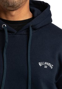 Sudadera color azul marino hecha de una mezcla de algodón que presenta una capucha con cordón, textura gruesa y un logotipo bordado en blanco de "BILLABONG" en la parte frontal.