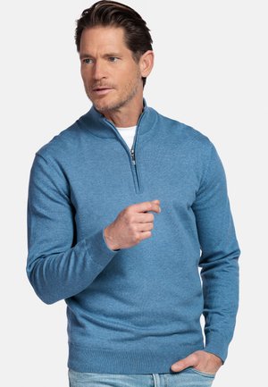 Man draagt een lichtblauwe trui met een korte rits en een spijkerbroek, met één hand in zijn zak en de andere hand geheven nabij zijn borst.