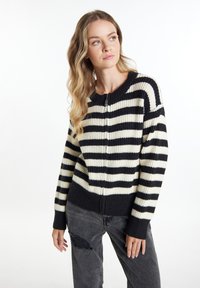 Junge Frau mit langen blonden Haaren, die einen schwarz-weißen gestreiften Reißverschluss-Pullover und dunkle zerrissene Jeans trägt und zur Seite schaut.