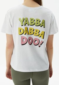 Camiseta de algodón blanca con texto audaz y colorido en la parte posterior: "YABBA DABBA DOO!" en verde, rosa y negro. Corte holgado, mangas cortas.