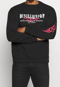 Sudadera negra con cuello redondo. Presenta un llamativo texto gráfico en blanco y rosa en la parte delantera y un diseño en rosa en la manga. La tela parece suave y texturizada.