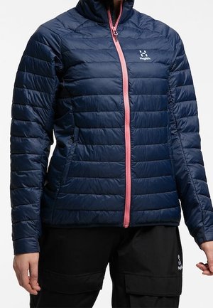 Mujer con chaqueta acolchada azul marino con cremallera rosa y logo en el pecho, combinada con pantalones cargo negros.