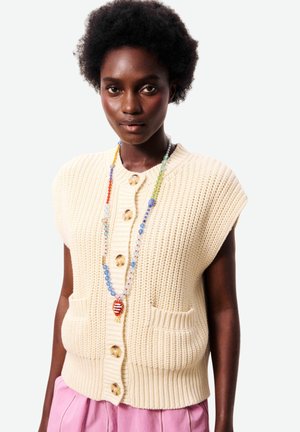 Femme portant un gilet sans manches crème en tricot avec de gros boutons, un pantalon rose et un long collier multicolore de perles.