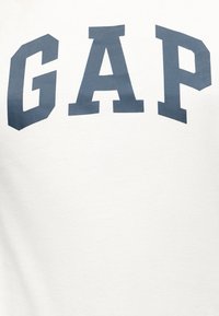 Tissu blanc avec de grandes lettres majuscules bleu foncé formant le mot "GAP" au centre supérieur.