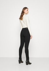 Black skinny jeans with a high waist, paired with a cream turtleneck top. The model wears black heeled boots, showcasing a fitted silhouette.