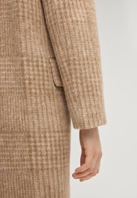 Beigefarbener und brauner kariert Wollmantelärmel und Seitentasche mit einer entspannt nach unten ausgestreckten Hand vor neutralem Hintergrund.