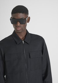 Giacca nera con zip e due tasche frontali, realizzata in tessuto texturizzato. Abbinata a occhiali da sole neri oversize, con montatura angolare.