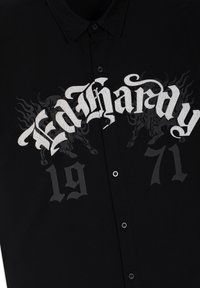 Černá košile na knoflíky s bílým textem ve staroanglickém stylu „Ed Hardy“ přes hruď a slabým tmavým motivem dvou hořících býků za textem.
