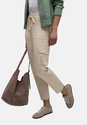 Personne portant un pantalon cargo beige, une veste vert clair, des baskets beige, et tenant un grand sac à bandoulière en daim marron.