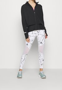 Persona vestita con giacca nera con zip, leggings bianchi con motivo astratto nero e pannelli in rete, e sneakers azzurro chiaro, in piedi su uno sfondo semplice.