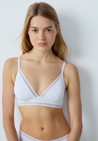 Bralette blanca con forma triangular, delicado borde de encaje, tirantes ajustables y textura de tela suave. Diseño simple y minimalista.
