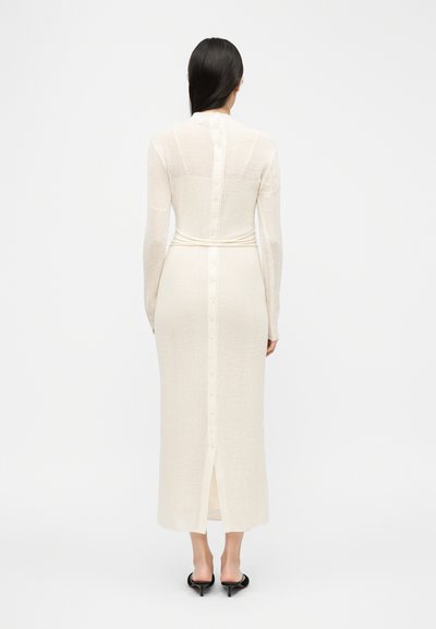 Femme portant une longue robe crème côtelée avec des boutons dans le dos, associée à des mules à talons noires, debout de dos.