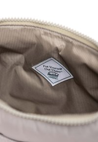 Sisäkuva laukusta, jossa on vaaleanbeige kangaspinnoite, jossa on vaaleita raidoja, sekä valkoinen etiketti, jossa lukee "Asetu itsesi esille™" merkiltä Herschel.