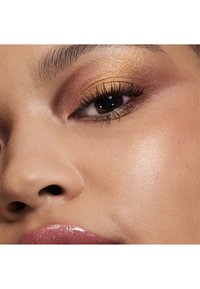 Gros plan sur un œil maquillé avec des ombres à paupières dorées et marron, de longs cils, et une peau éclatante à côté d'une lèvre brillante avec une subtile teinte rose.