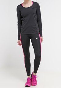 Musta pitkähihainen urheiluvaate yhdistettynä mustiin legginsseihin, joissa on vaaleanpunaiset sivuviivat, sekä kirkkaat vaaleanpunaiset urheilukengät. Nike-logot näkyvissä.