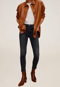 Person trägt eine braune Lederjacke mit Knöpfen, weißes T-Shirt, schwarze eng anliegende Jeans und braune Knöchelstiefel, die Hände in den Taschen.