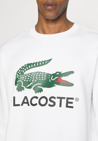 Fehér kapucnis pulóver, zöld krokodil logóval, piros részletekkel és fekete "LACOSTE" felirattal. Pamut keverék anyag, sima tapintású.