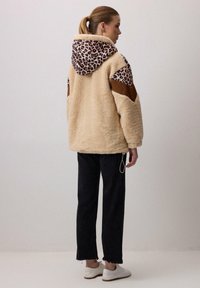 Chaqueta de pelo sintético beige con capucha de estampado de leopardo, detalles en marrón en los hombros, corte holgado y cordón elástico en el dobladillo. Combinada con pantalones negros.