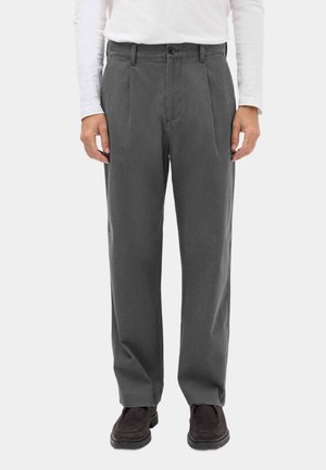 Man draagt donkergrijze geplooide broek, witte top met lange mouwen en donkerbruine schoenen, staand tegen een effen achtergrond.