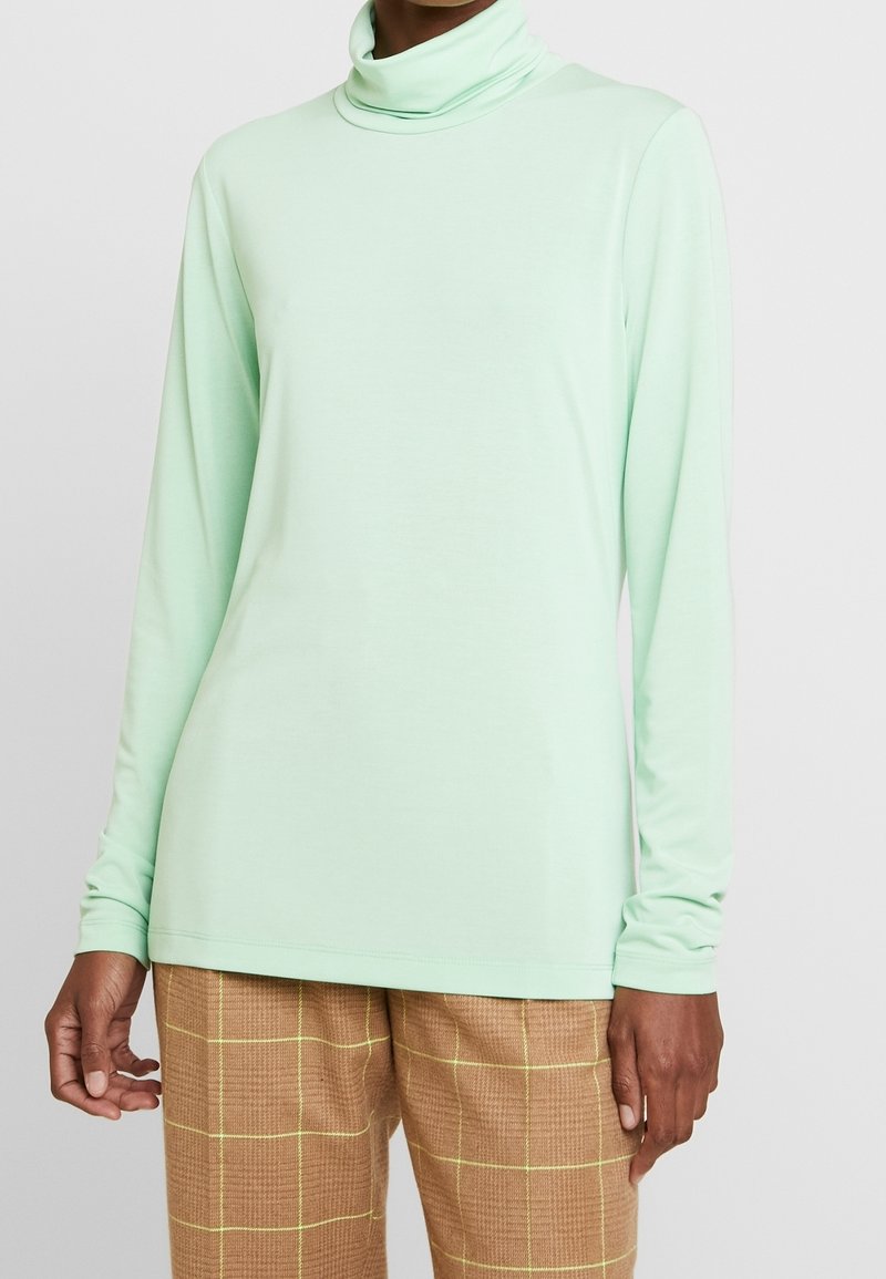 Lättgrön långärmad tröja med hög krage, tillverkad av mjukt material; har en avslappnad passform och en slät yta. Matchas med bruna rutiga byxor.