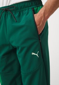 Gröna sportkortbyxor i lätt material, med dragkedjefickor, elastisk midja och en reflekterande vit Puma-logotyp på låret.