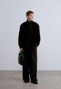 Μαύρο μπουφάν με ψηλό γιακά, φορεμένο πάνω από μαύρο ζιβάγκο. Φαρδιά μαύρη παντελόνια και γυαλιστερά μαύρα παπούτσια. Κρατάει μαύρη δερμάτινη τσάντα.