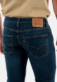 Jeans in denim blu scuro con un classico design a cinque tasche. Presentano una toppa posteriore in pelle marrone con il logo del marchio e dettagli di cucitura.