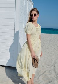 Gul gingham-maxiklänning med puffärmar och resår i midjan. Kombinerad med bruna flip-flops, placerad framför en strand och en vit byggnad.