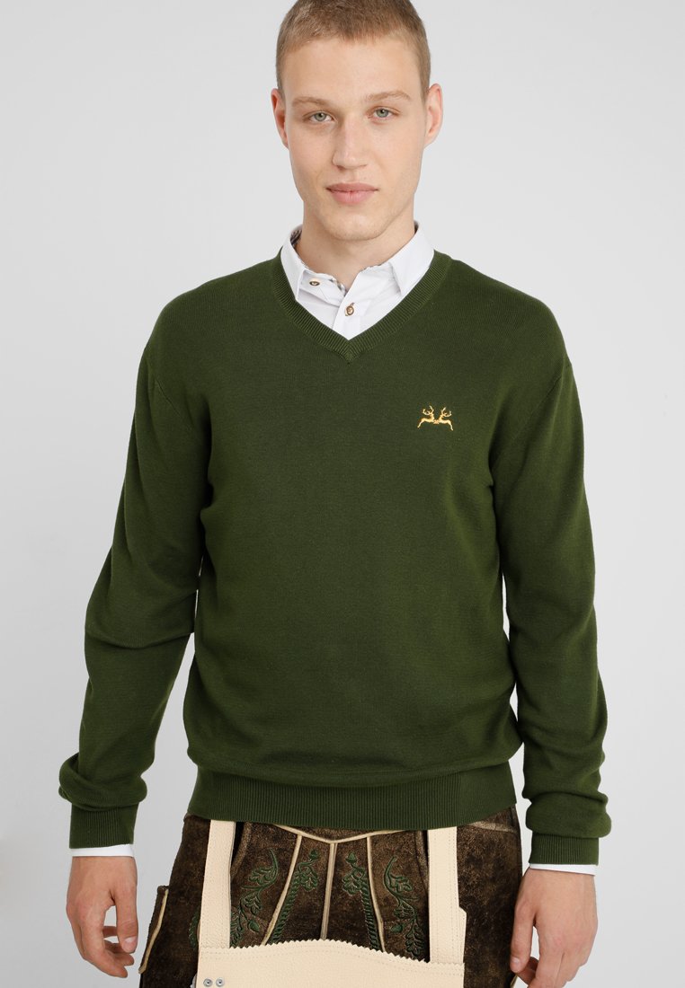 Grüner Wollpullover mit V-Ausschnitt, der ein goldenes besticktes Design auf der linken Brust zeigt, kombiniert mit einem weißen Hemd darunter.