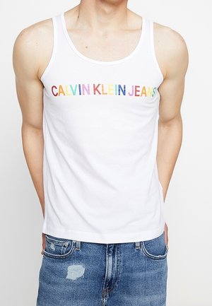 Biały bezrękawnik z wielokolorowym napisem "CALVIN KLEIN JEANS" noszony z niebieskimi, postarzanymi jeansami.