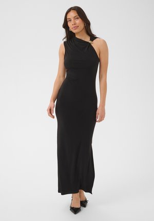 Femme debout portant une robe longue noire moulante, sans manches, à encolure asymétrique et chaussures noires pointues, sur un fond uni.