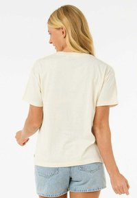 Camiseta de algodón color crema con mangas cortas y un corte relajado. La parte trasera presenta un diseño liso, complementando unos pantalones cortos de mezclilla claros.