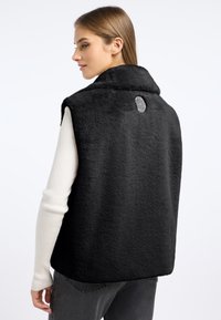 Czarna bezrękawnik wykonana z miękkiego, puszystego materiału z wysokim kołnierzem i dekoracyjną metką z tyłu. Noszona na lekkim swetrze.