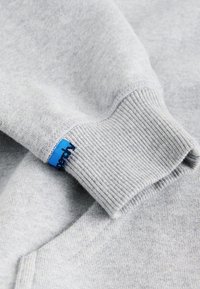 Hellgrauer Sweatshirt-Ärmel mit geripptem Bündchen und einem kleinen blauen Stoffetikett nahe der Handgelenksnaht.