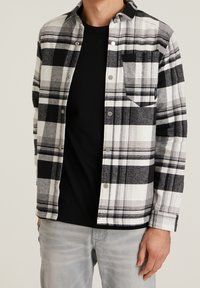 Zwart-wit geblokt overhemd met knopen en een borstzak, lange mouwen en grijze stof eronder. De knopen zijn van zilverkleurig metaal.
