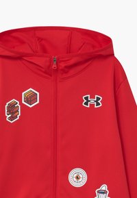 Punainen vetoketjuhuppari, joka on valmistettu sileästä kankaasta ja jossa on huppu. Hupparissa on useita värikkäitä grafiikoita ja etuosassa Under Armour -logo.