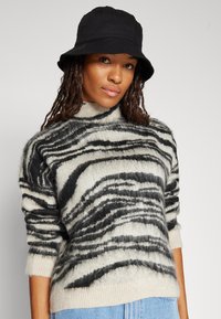 Gestreifter Pullover in Creme und Schwarz aus weichem, strukturiertem Stoff mit hohem Kragen und weiten Ärmeln, kombiniert mit einem schwarzen Bucket Hat.