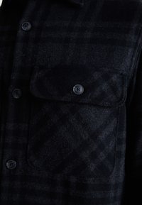 Cămașă din flanel neagră cu un model în carouri, având un buzunar pe piept și butoane negre. Textura pare moale și grea.