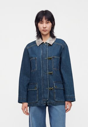 Jeune personne aux cheveux noirs portant une veste en jean bleu foncé avec un col à carreaux et des fermetures à crochets métalliques, assortie d’un jean bleu clair.