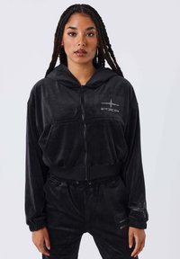 Sudadera corta de terciopelo negro con cierre de cremallera, bolsillos frontales, dobladillo acanalado y diseño de logo de estrella. Combinada con pantalones jogger de terciopelo a juego.