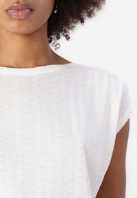 White top with textured fabric, wide neckline, and short sleeves, featuring a subtle pattern of small rectangular shapes.