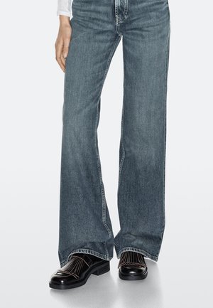 Blå jeans med vide ben kombineret med sorte lædersko med plisserede detaljer og lave hæle, på en ensartet lys baggrund.