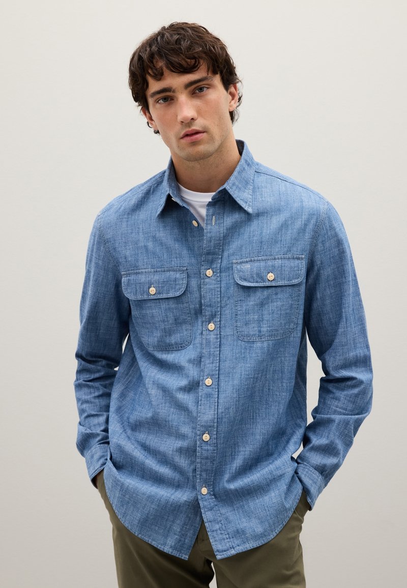 Camisa de chambray azul claro con mangas largas, dos bolsillos en el pecho y cierre de botones. La tela tiene un aspecto texturizado y ligeramente deslavado.