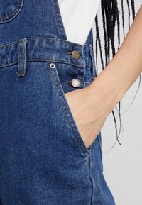 Käsi, jolla on vaalea ihonväri, lepäilee sinisten farkkuhaalareiden etutaskussa, jotka on puettu valkoisen paidan päälle.