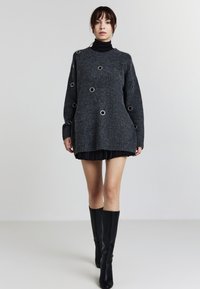Maglione oversize grigio scuro lavorato a maglia con ritagli circolari e occhielli argentati, abbinato a una gonna nera plissettata e stivali neri al ginocchio.