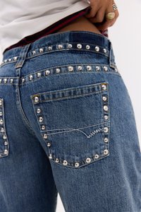 Close-up of blue denim jeans with silver studs outlining the back pockets and waistband, worn with a partially lifted white shirt.
